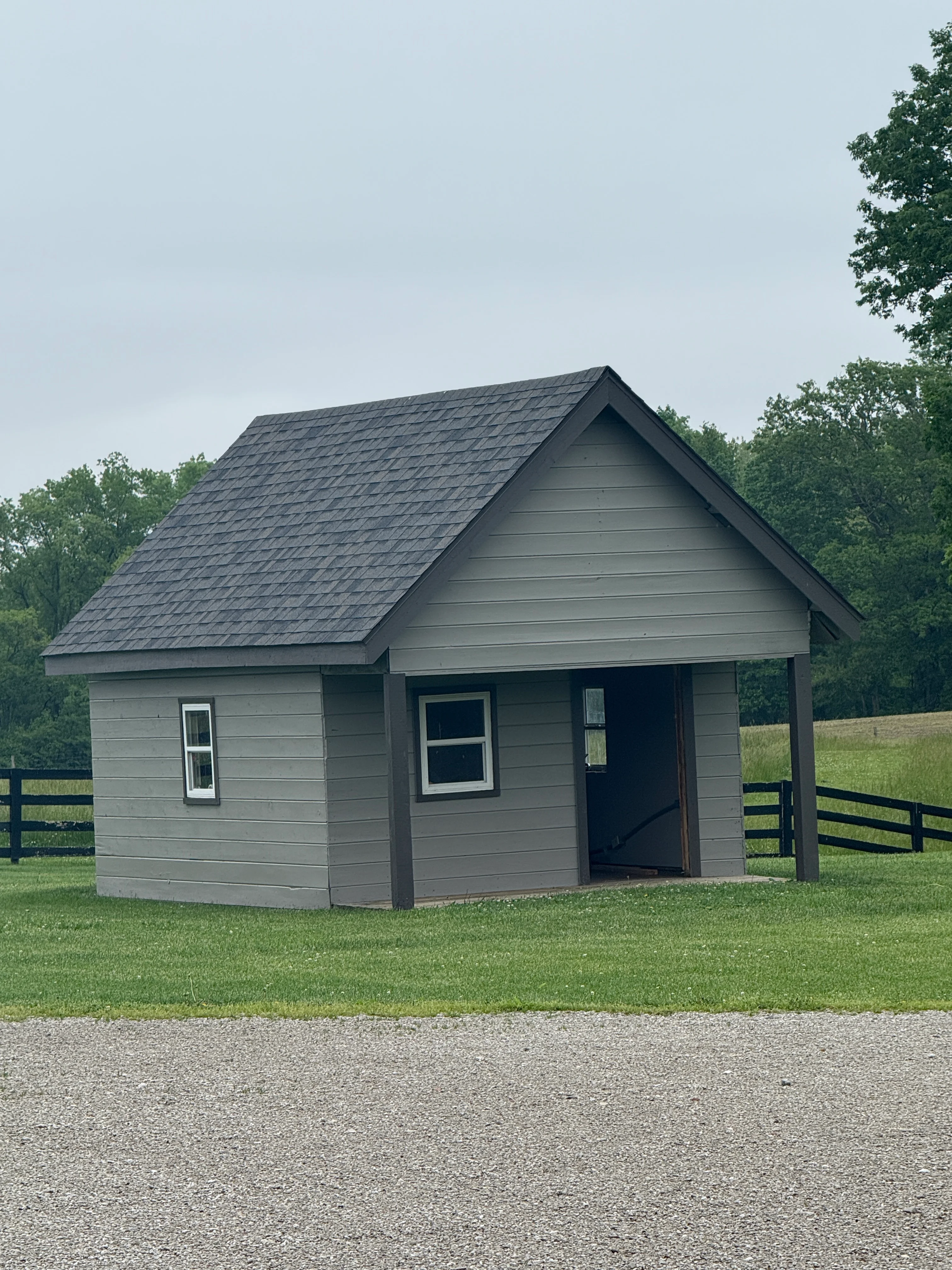 Gray painted shed with white trim windows, dark roof, and covered porch on grassy property
