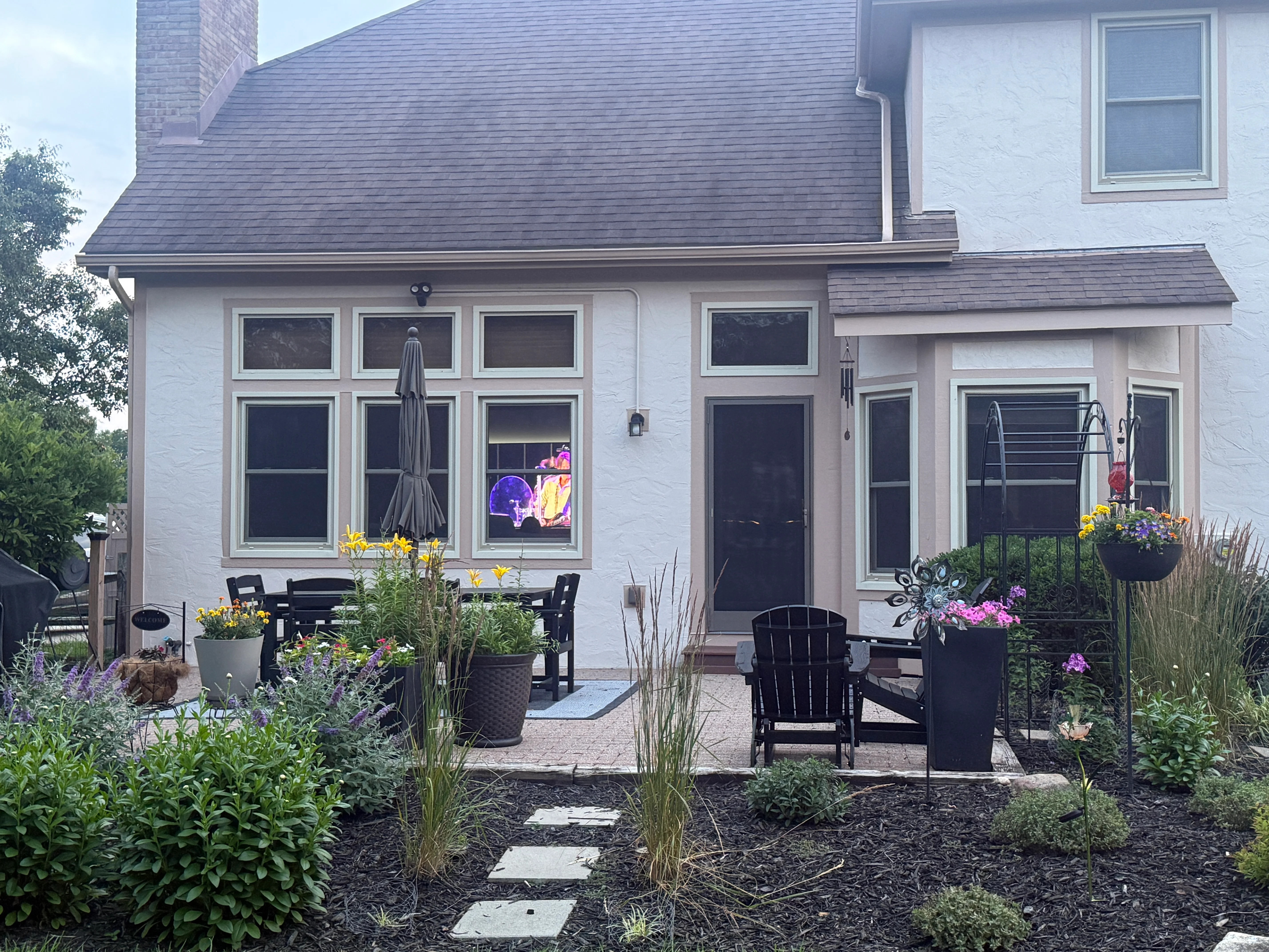 White farmhouse with dark-trimmed windows, slate roof, and manicured patio garden with black Adirondack chairs